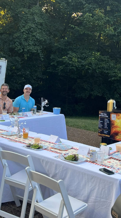Outdoor birthday party with friends seated around a long white-tableclothed dining table, plated salads and drinks, a food cart and trailer against a wooded backyard backdrop.