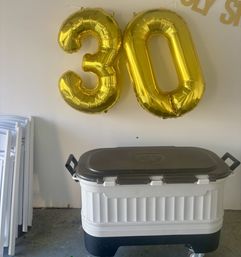 Shiny gold foil '30' balloons floating above a white wheeled cooler and stacked folding chairs against a plain wall — casual indoor 30th birthday party setup.