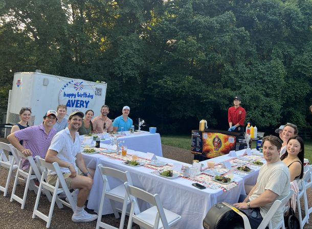 Outdoor birthday party for Avery — friends seated at a long white-tablecloth table with salads and drinks, a chef at a food cart grilling, wooded backyard backdrop.