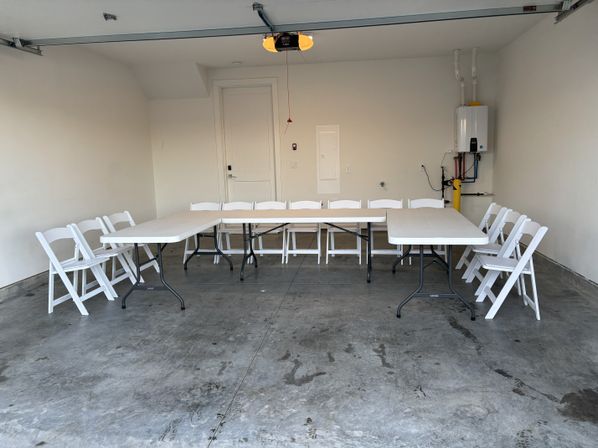 Empty home garage set up for a gathering: three white folding tables arranged in a U-shape with white folding chairs around them on a stained concrete floor, garage door opener overhead and a tankless water heater on the right wall.