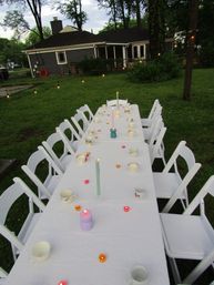 Backyard alfresco setup with a long white-tablecloth table and white folding chairs, scattered teacups and assorted lit candles, string lights overhead and a house in the background creating a cozy evening scene.