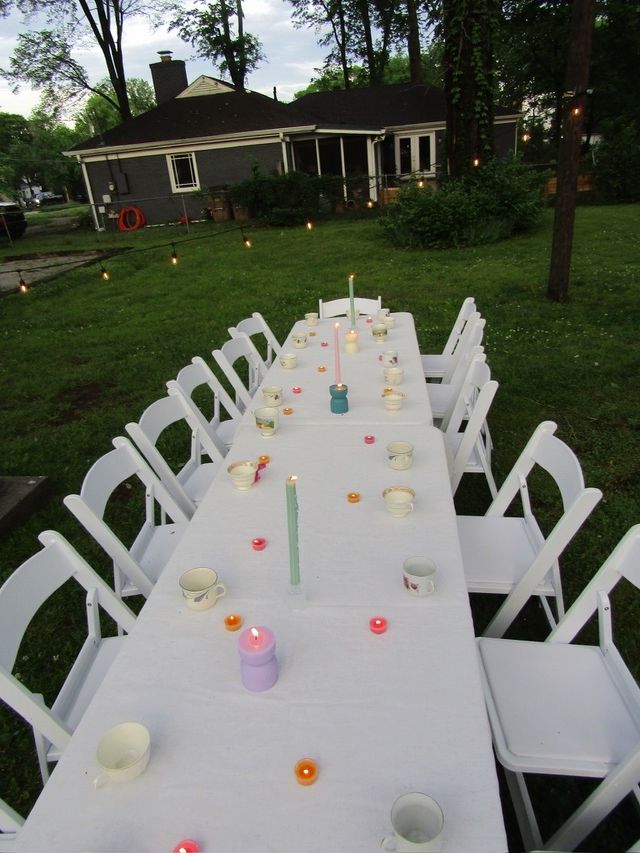 Backyard alfresco setup with a long white-tablecloth table and white folding chairs, scattered teacups and assorted lit candles, string lights overhead and a house in the background creating a cozy evening scene.