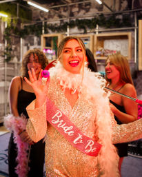 Smiling bride-to-be in a glittery gold dress and white feather boa wearing a pink "Bride to Be" sash, holding a small pink party token and laughing with friends at an indoor bachelorette party.