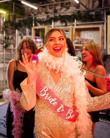 Smiling bride-to-be in a glittery gold dress and white feather boa wearing a pink "Bride to Be" sash, holding a small pink party token and laughing with friends at an indoor bachelorette party.