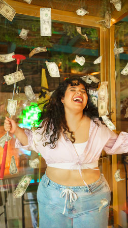 Laughing woman in an indoor cash-grab money booth with dollar bills swirling around her, holding a red handle, wearing a pink crop top and jeans
