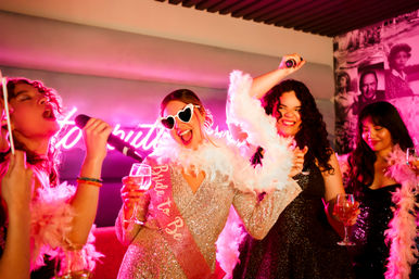 Bachelorette party karaoke — bride-to-be in heart sunglasses and feather boa singing with friends under pink neon lights, cocktails in hand