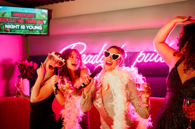 Bachelorette party women belting karaoke in a neon-lit karaoke bar, wearing heart sunglasses and a feather boa while holding cocktails — lively nightlife celebration.