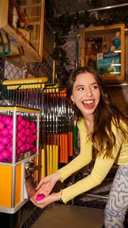 Smiling woman in a yellow top collecting a bright pink golf ball from a coin-operated dispenser at an indoor mini-golf arcade with colorful putters and games in the background.