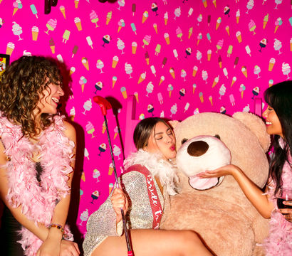 Three women in sparkly party outfits and pink feather boas pose in a playful pink room with ice-cream patterned wallpaper; one kisses a giant teddy bear while holding a red mini-golf putter as friends laugh and hold drinks.