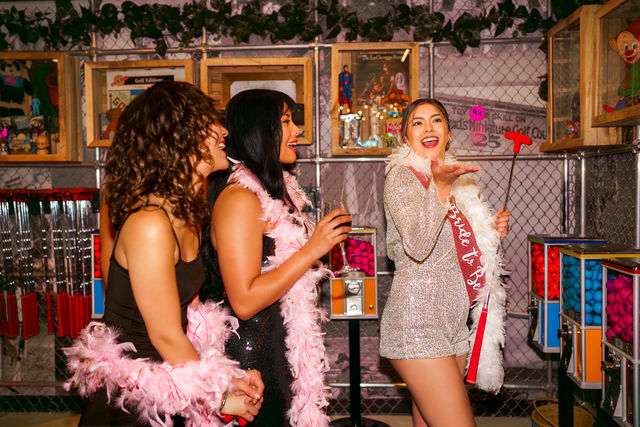 Three friends at a bachelorette party in a colorful indoor mini-golf arcade, bride-to-be in a sparkly dress with sash and feather boa blowing a kiss while friends cheer with drinks