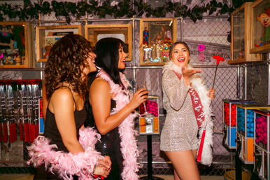 Bachelorette party at an indoor mini-golf arcade — smiling bride-to-be in a sequin outfit and sash swinging a mini putter while friends in pink feather boas cheer by colorful gumball machines