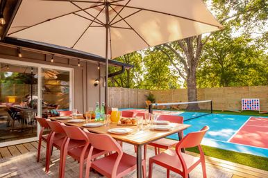 Sunlit backyard patio with umbrella-shaded wooden dining table set for six with red chairs, glassware and pitchers, adjacent to a blue-and-red outdoor sport court and fenced yard under mature trees.