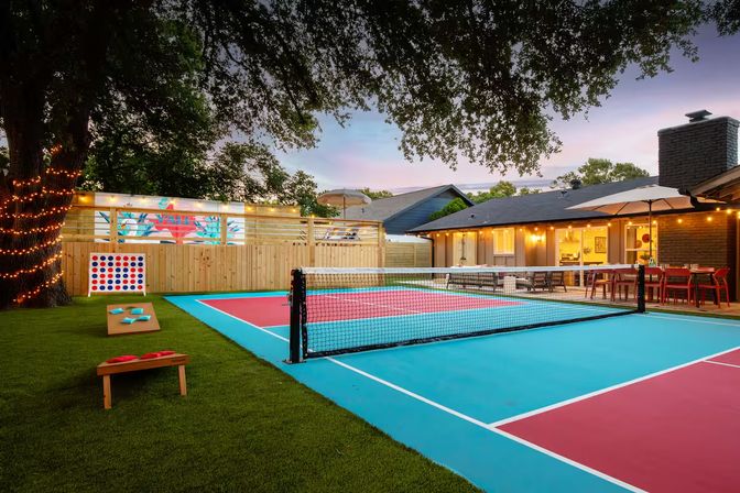 Vibrant suburban backyard pickleball court at dusk with turquoise and coral courts and net, cornhole boards and oversized Connect Four on green turf, a tree wrapped in string lights and an illuminated outdoor dining patio beside a single‑story house