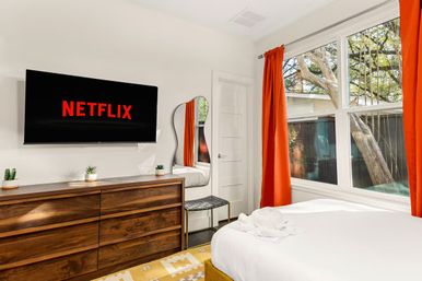 Bright modern bedroom with wall-mounted TV displaying Netflix, mid-century wooden dresser, white bed with folded towel, large window with orange curtains and backyard trees visible outside