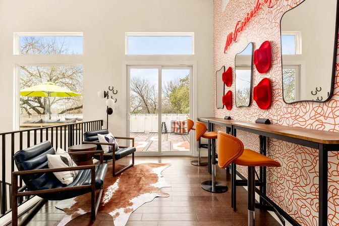 Sunny modern lounge with sliding glass doors to a tree-lined balcony, navy leather armchairs on a cowhide rug, long wooden counter with orange bar stools and sculptural red wall hooks and mirrors.