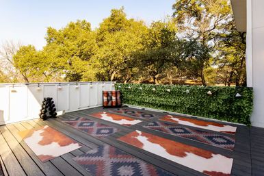 Sunlit outdoor yoga/fitness deck with southwestern and cowhide-style mats on dark wood planks, ivy-covered railing with string lights, stacked weights, and oak trees beyond.