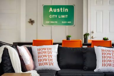 Cozy Austin living room with a black sofa and white throw pillows reading HOWDY repeatedly, bright orange dining chairs at a table with small potted plants, and a green Austin City Limit sign on the wall.