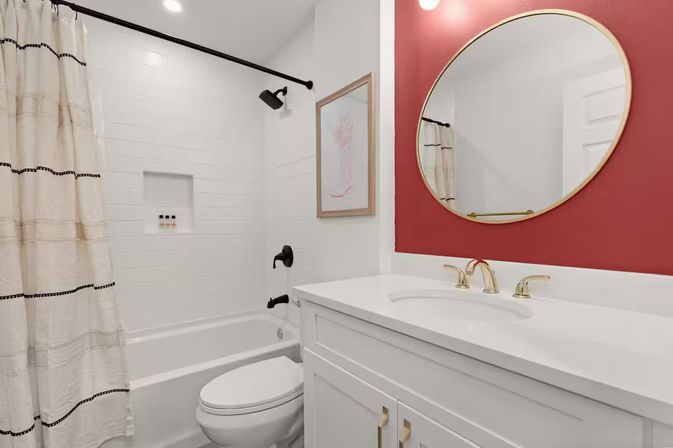 Bright modern bathroom with white subway-tiled bathtub and shower, cream patterned curtain, white toilet and vanity with gold faucets, and a round mirror on a red accent wall