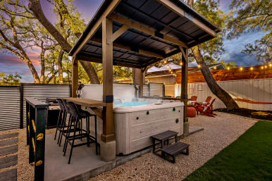 Backyard covered hot tub under a wooden pergola with bar seating, string lights, Adirondack chairs, hammock and oak trees at sunset