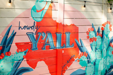 Colorful mural on a wooden fence reading “howdy Y’ALL” with a red Texas silhouette, turquoise cacti, cowboy hat and string lights — Austin, Texas vibe.