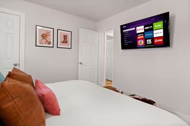 Cozy modern bedroom with white walls, wall-mounted smart TV displaying streaming apps, white bed topped with brown leather and pink pillows, two framed prints and an open door to a hallway.