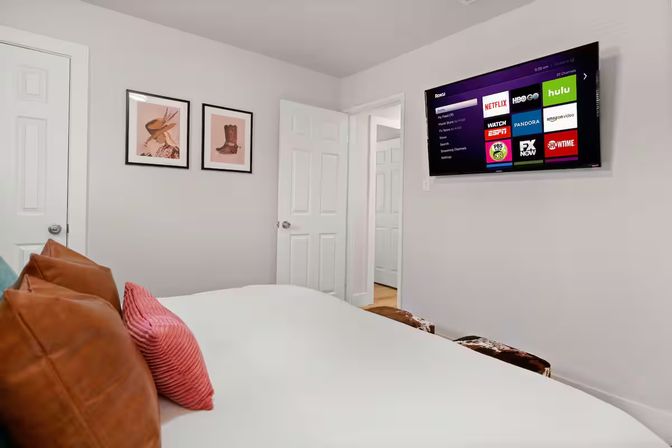 Cozy modern bedroom with white walls, wall-mounted smart TV displaying streaming apps, white bed topped with brown leather and pink pillows, two framed prints and an open door to a hallway.