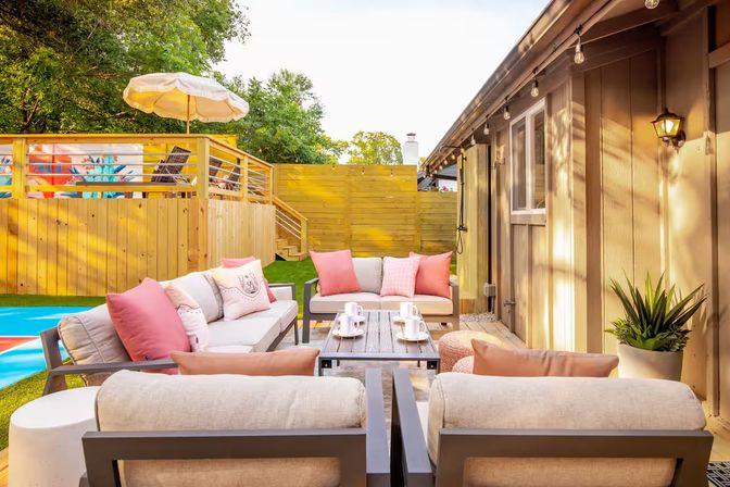 Sunny backyard patio outdoor seating area with modern cushioned sofas and coral throw pillows around a coffee table, wooden deck, umbrella and string lights.
