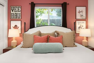 Texas-inspired bedroom with coral accent wall, white upholstered headboard, beige and coral throw pillows and mint bolster, bedside lamps, 'YEE HAW' wall art and a window framed by dark curtains.