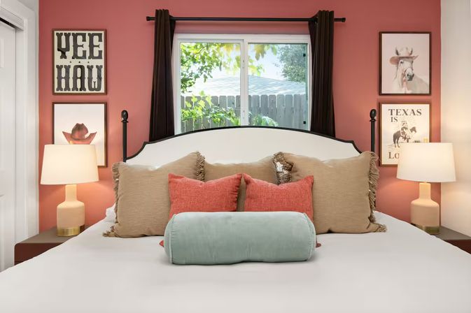 Texas-inspired bedroom with coral accent wall, white upholstered headboard, beige and coral throw pillows and mint bolster, bedside lamps, 'YEE HAW' wall art and a window framed by dark curtains.
