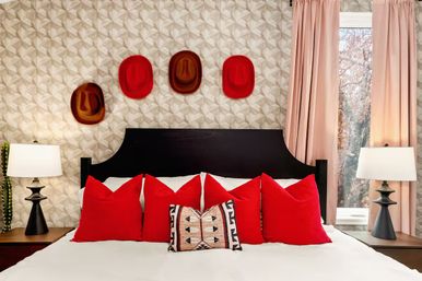 Modern cozy bedroom with black headboard and geometric wallpaper, playful row of four hats mounted above, white bedding styled with five vibrant red throw pillows and a patterned lumbar pillow, flanked by matching bedside lamps and soft pink curtains.
