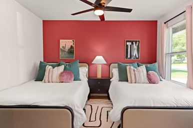 Sunny twin bedroom with a red accent wall, two matching beds with white linens, teal and blush throw pillows, a central nightstand and lamp, ceiling fan, patterned rug and a large window — modern playful decor with Austin/TX wall art.
