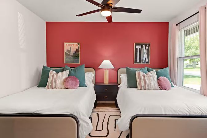 Sunny twin bedroom with a red accent wall, two matching beds with white linens, teal and blush throw pillows, a central nightstand and lamp, ceiling fan, patterned rug and a large window — modern playful decor with Austin/TX wall art.