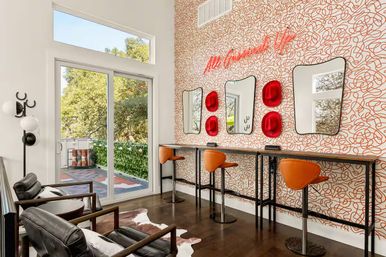 Sunlit modern lounge with three orange bar stools at a high wood counter against playful red-scribble wallpaper with irregular mirrors and red hat accents, black leather armchairs, cowhide rug, and sliding glass door opening to a leafy outdoor patio.