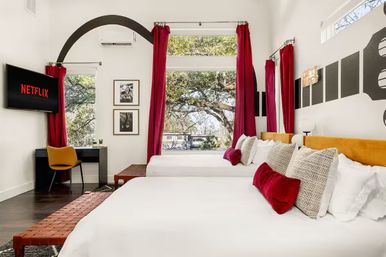 Bright modern boutique hotel room with two queen beds, red velvet pillows and curtains, large windows framing oak trees, desk workspace and Netflix-ready wall TV.