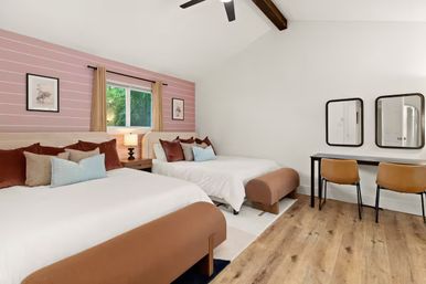 Bright modern guest room with two beds in white linens, rust and blue throw pillows, pink striped accent wall, window with tan curtains, bedside lamp, vaulted ceiling with exposed wood beam and ceiling fan, hardwood floors, and a desk with two leather chairs and mirrors.
