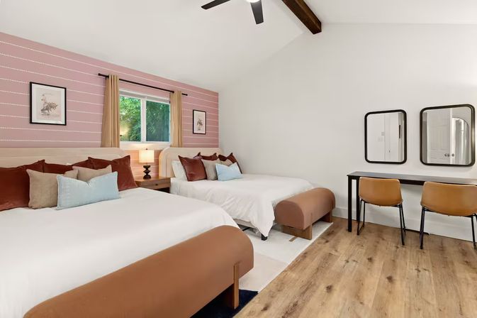 Bright modern guest room with two beds in white linens, rust and blue throw pillows, pink striped accent wall, window with tan curtains, bedside lamp, vaulted ceiling with exposed wood beam and ceiling fan, hardwood floors, and a desk with two leather chairs and mirrors.