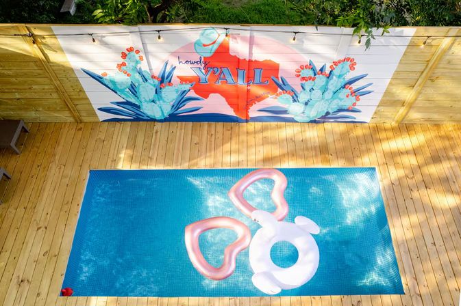 Aerial view of a backyard rectangular pool with pink heart-shaped and white donut inflatables on blue water, surrounded by a wooden deck and a colorful Texas silhouette and cactus mural reading “Howdy Y’all”.