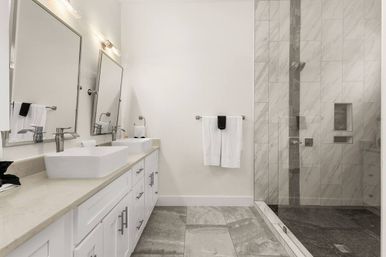 Bright modern master bathroom with white double vessel sinks, large mirrors, gray stone tile floor, and glass-enclosed marble walk-in shower with white towels