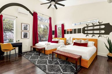 Sunlit modern music-themed bedroom with two orange upholstered beds dressed in white linens and red accent pillows, guitar headstock wall decal, dark hardwood floors with a black-and-white geometric rug, woven benches, red curtains framing large windows overlooking trees, yellow desk chair and ceiling fan.