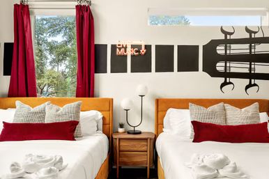 Boutique hotel room with two beds with mustard headboards, red curtains, guitar wall mural and a glowing "Live Music" neon sign above a wooden nightstand and modern lamp.