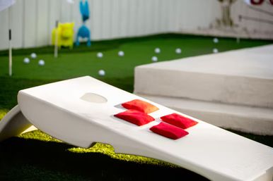 White cornhole board with four red bean bags on a green lawn, putting green and scattered golf balls in the background