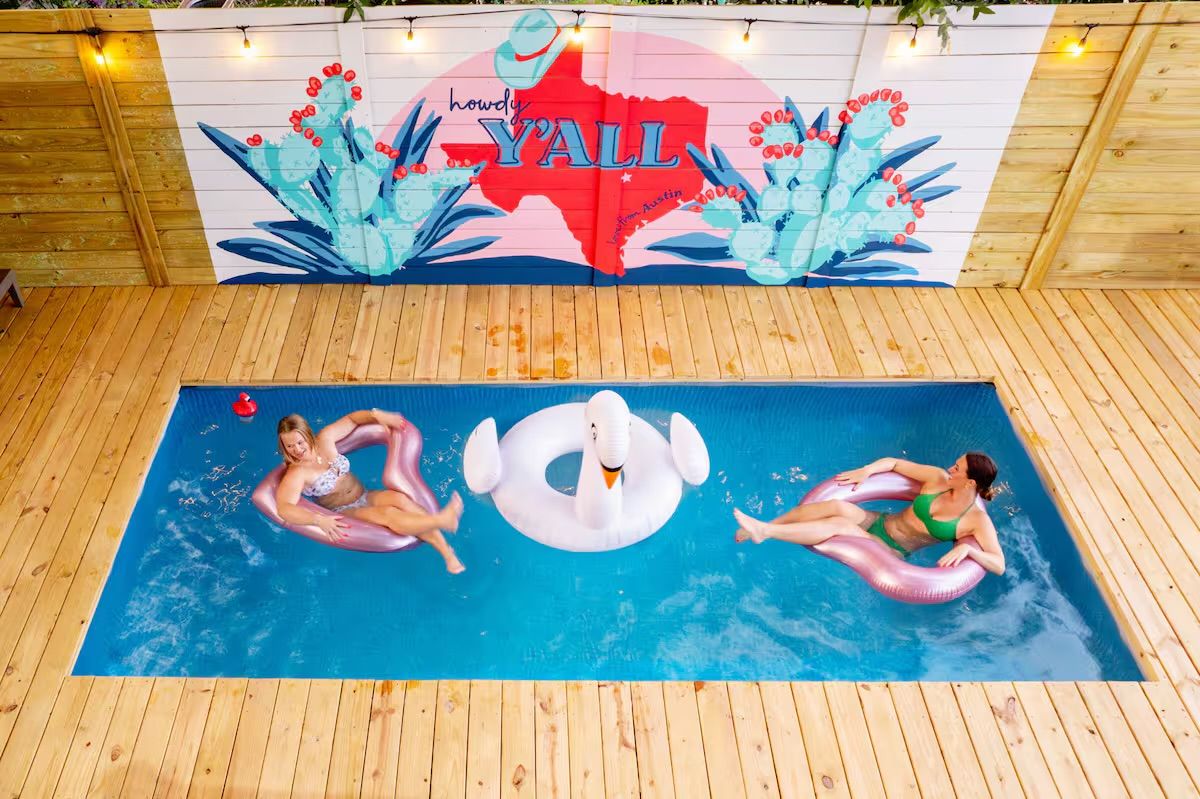 Aerial view of a small rectangular backyard pool with wood decking, two people lounging on pink inflatable rings and a white swan float beneath a colorful “Howdy Y'ALL” Texas-themed mural