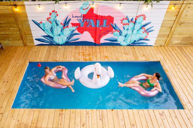 Aerial view of a small rectangular backyard pool with wood decking, two people lounging on pink inflatable rings and a white swan float beneath a colorful “Howdy Y'ALL” Texas-themed mural
