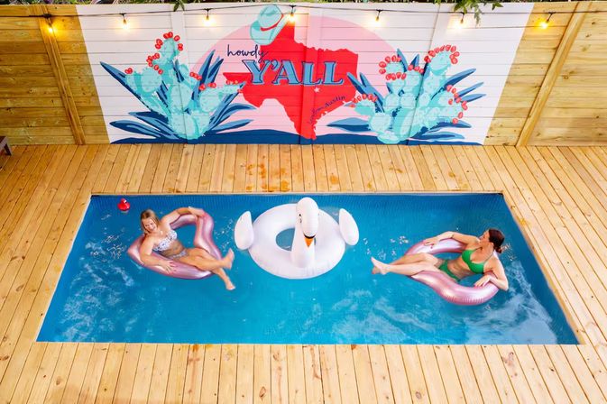 Aerial view of a small rectangular backyard pool with wood decking, two people lounging on pink inflatable rings and a white swan float beneath a colorful “Howdy Y'ALL” Texas-themed mural
