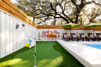 Poolside backyard with a small putting green and scattered golf balls, row of lounge chairs with patterned pillows under green umbrellas, white wood and cedar fence featuring a 'Long Live Cowgirls' mural, and large oak trees overhead.