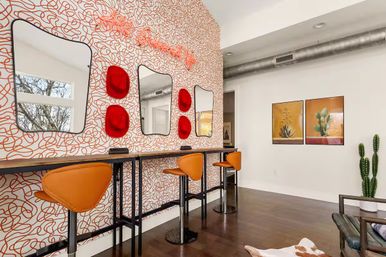 Contemporary studio interior with orange swivel bar stools at a long vanity counter, three curvy mirrors on orange-squiggle wallpaper, red wall accents, wood floors and a potted cactus.