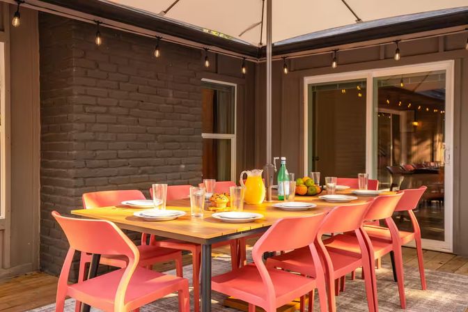 Sunlit backyard patio dining area with a wooden table and eight coral-colored chairs under a large umbrella and string lights, set with plates, glasses, a pitcher of orange juice, a bottle of sparkling water and a fruit bowl in front of sliding glass doors.