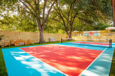 Vibrant red-and-turquoise outdoor pickleball court in a shaded fenced backyard, surrounded by large oak trees, Adirondack chairs and yard games.