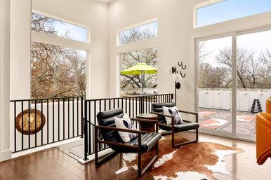 Sunlit contemporary living nook with two black leather armchairs and a small round table on a cowhide rug, tall windows and sliding glass door opening to a balcony with a lime umbrella and bare trees outside.