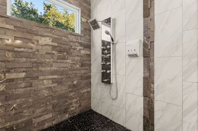 Modern indoor bathroom shower with stainless steel multi-jet panel, rainfall showerhead and handheld, white marble wall tiles, textured brown stone accent wall, black pebble floor and narrow window showing trees — spa-like design.
